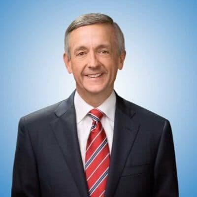 Smiling man with gray hair in a dark suit and red striped tie, blue background.