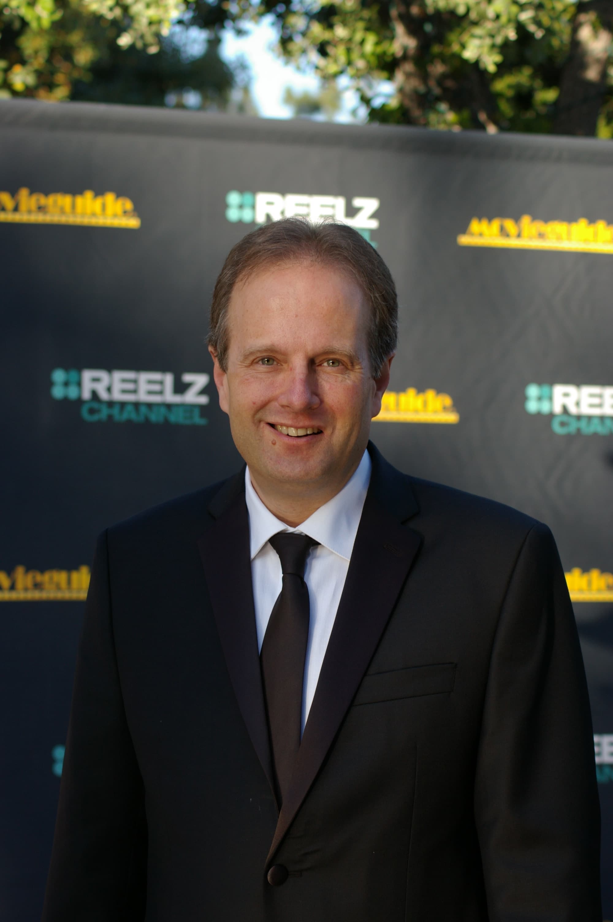 Smiling man in a black suit and tie posing against a Reelz Channel backdrop.