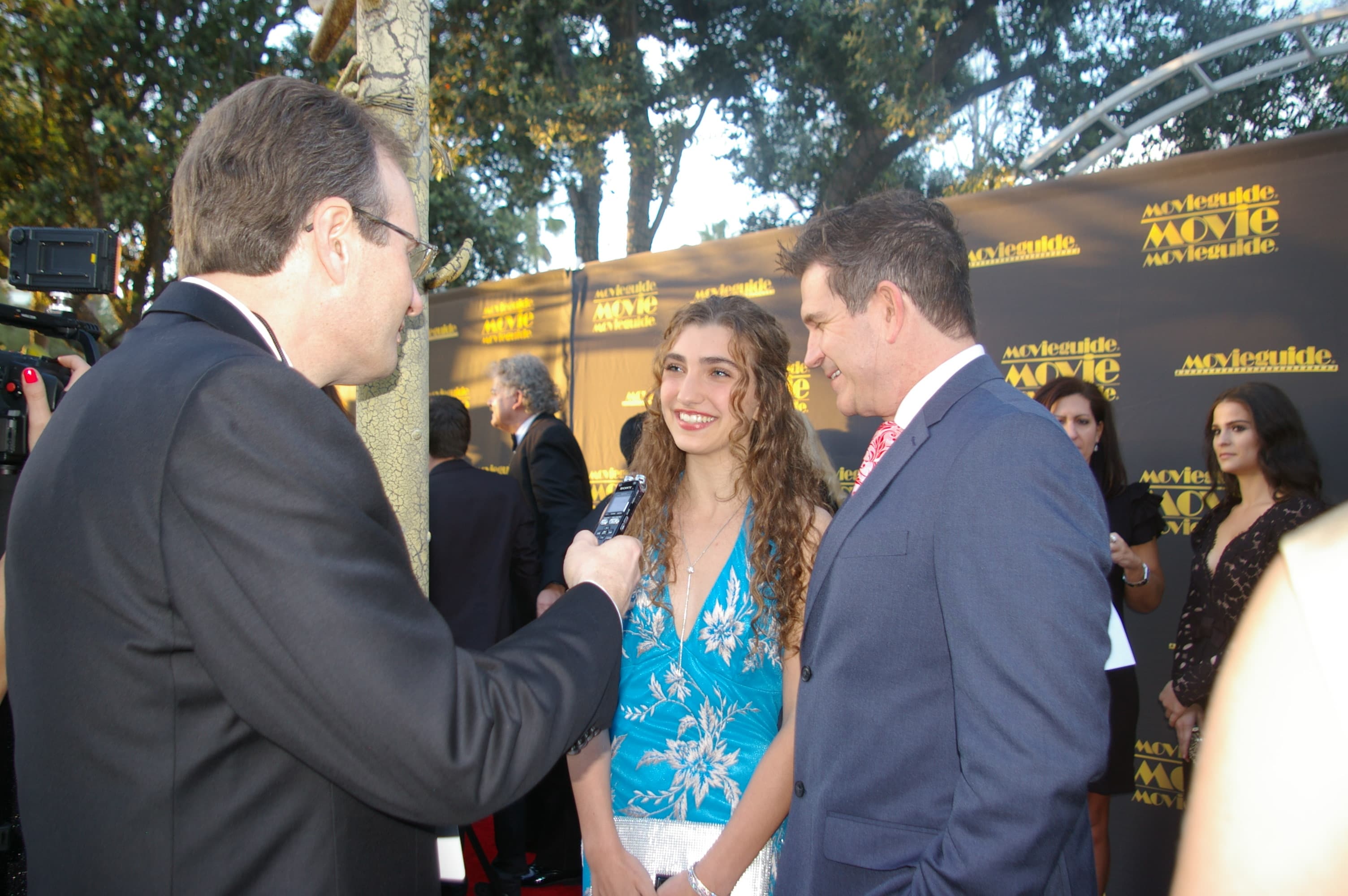 Reporter interviews a young woman in a blue dress and a man on red carpet.