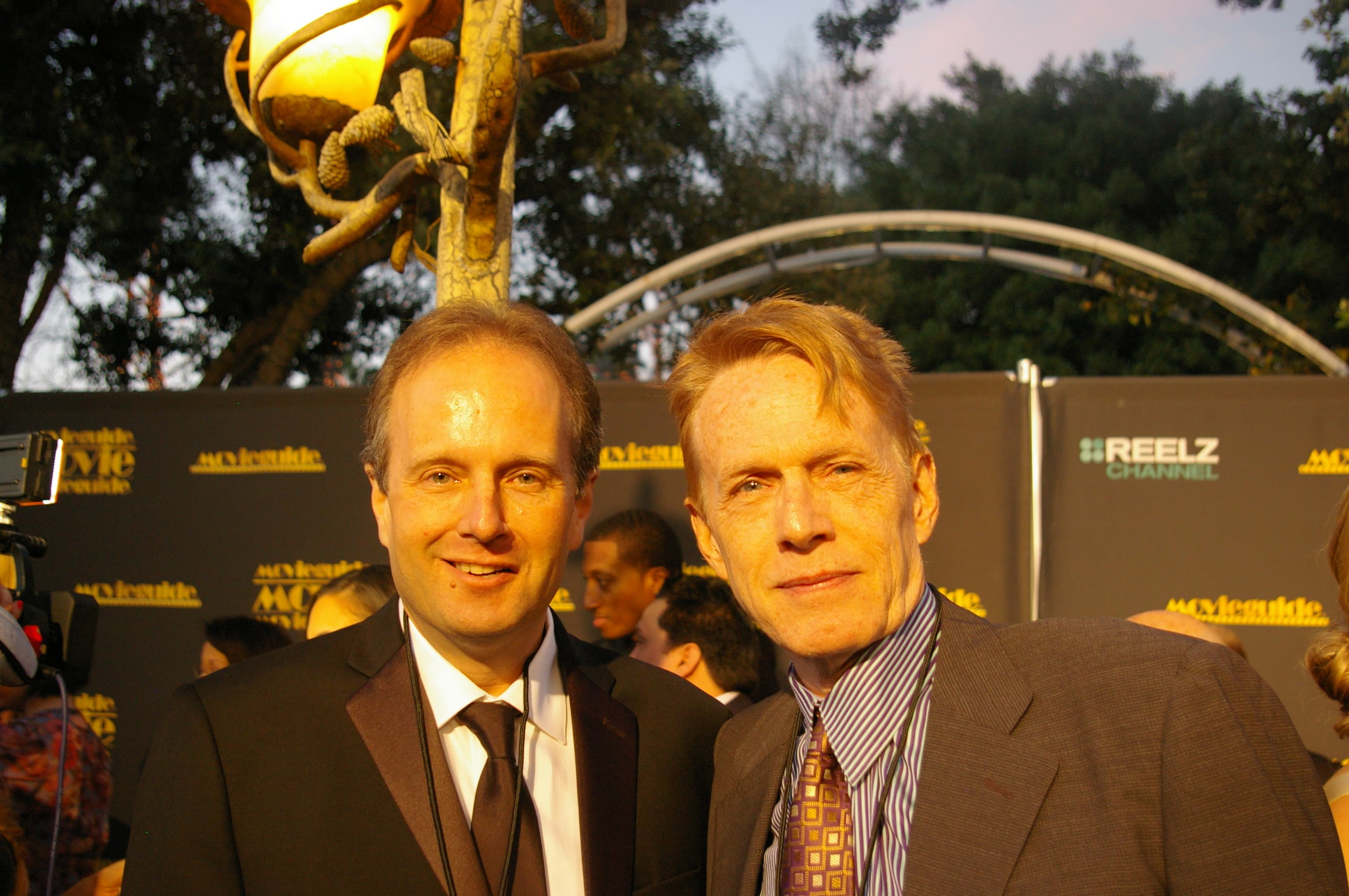 Two men in formal suits pose together at an outdoor red carpet awards event.