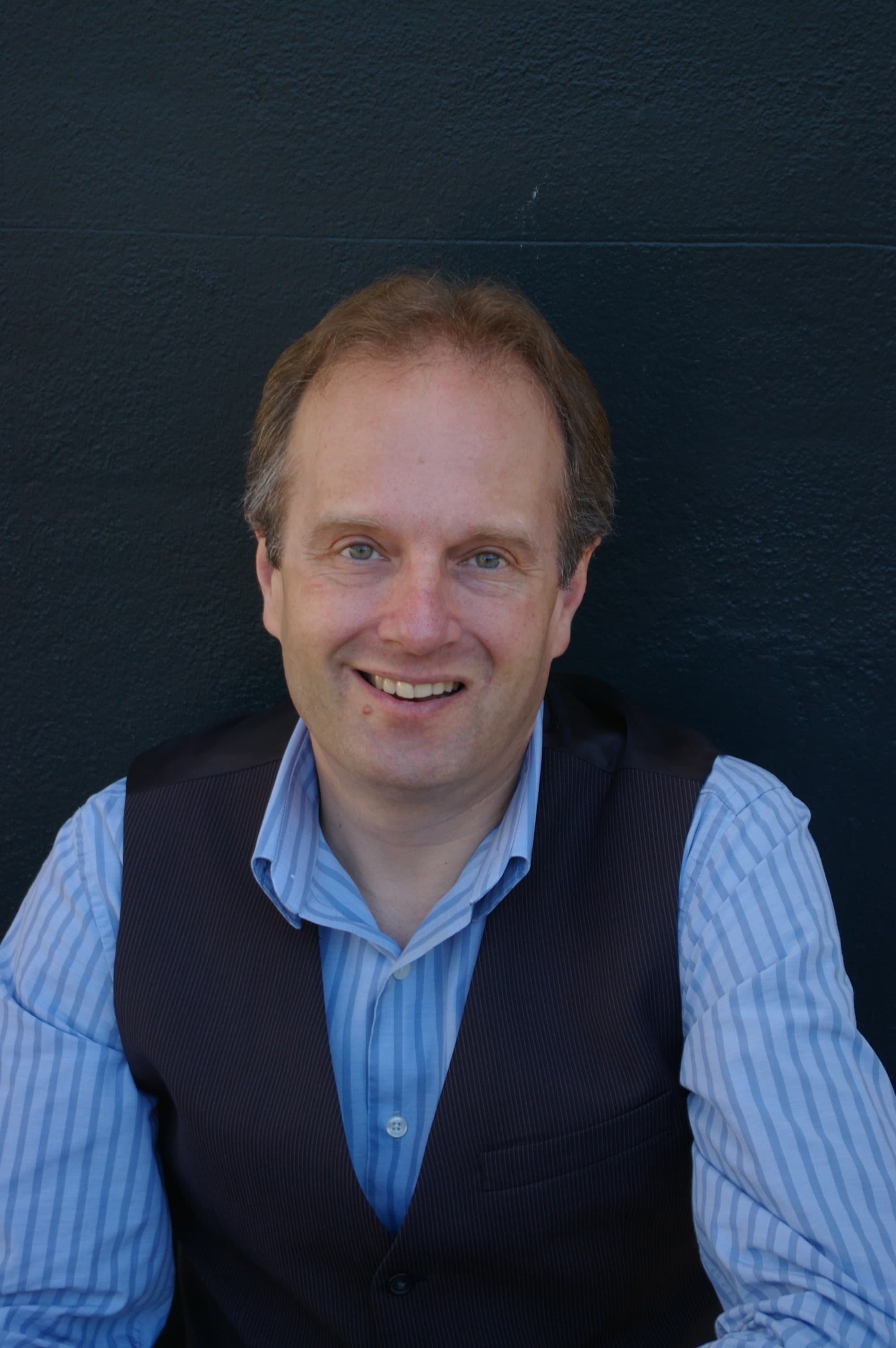 Smiling man with light brown hair wearing a blue striped shirt and dark pinstriped vest.