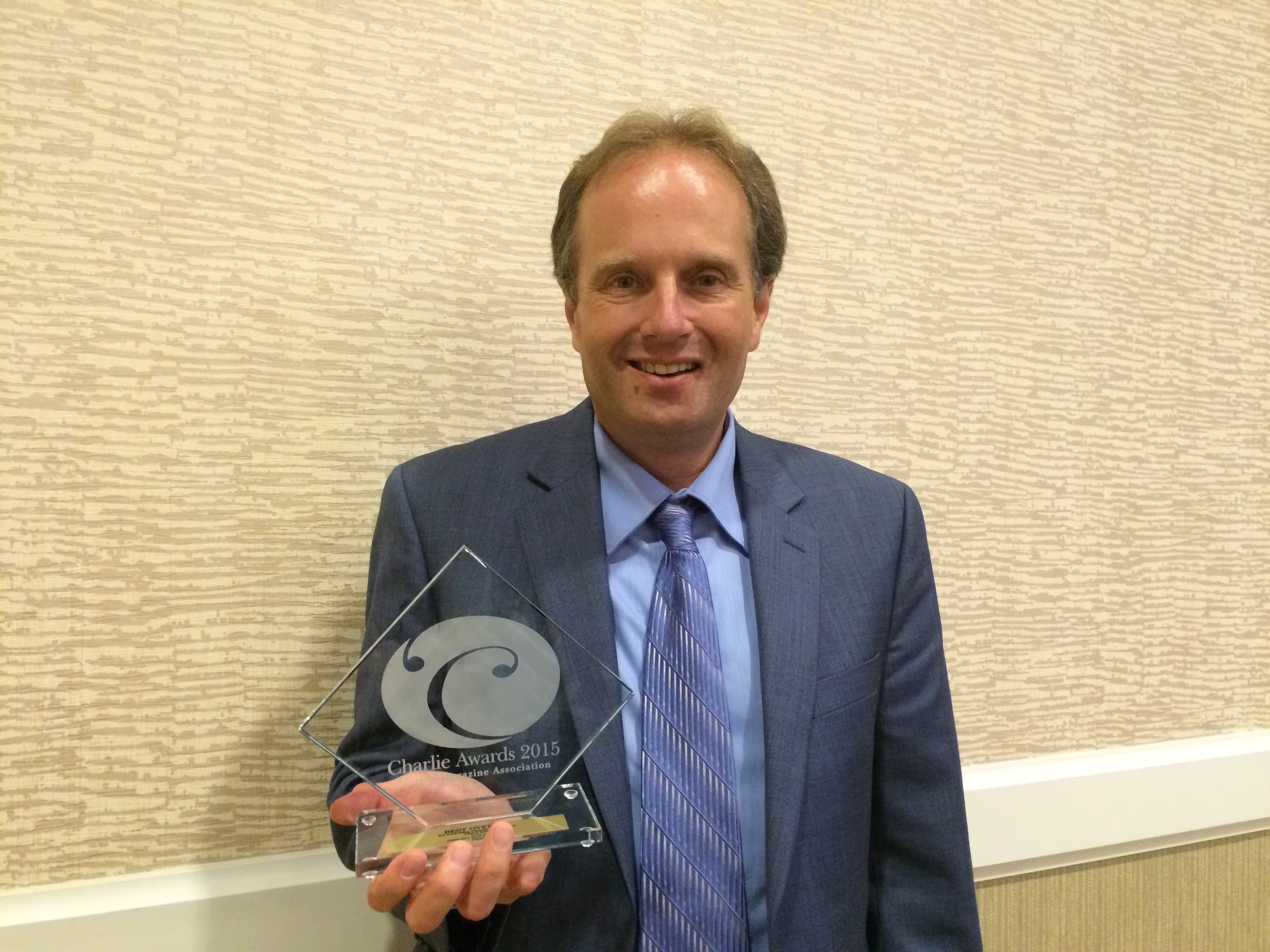 Smiling man in a blue suit holding a diamond-shaped glass Charlie Awards 2015 trophy.