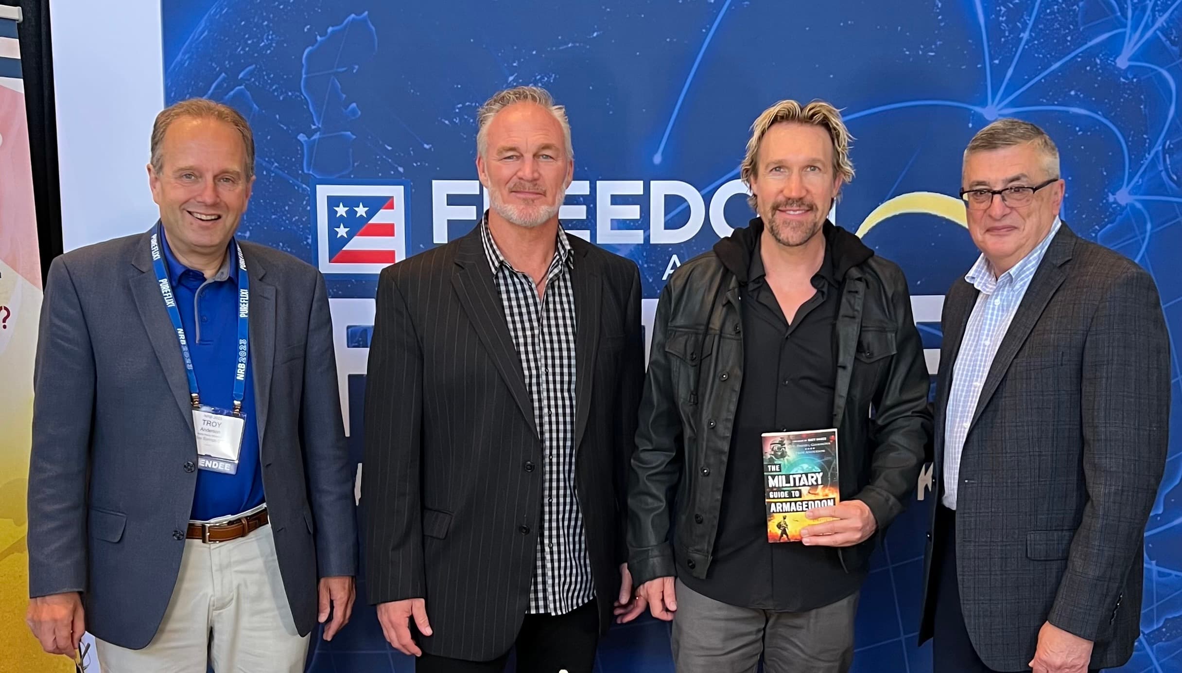 Four men pose before a blue Freedom banner, one holding The Military Guide to Armageddon.