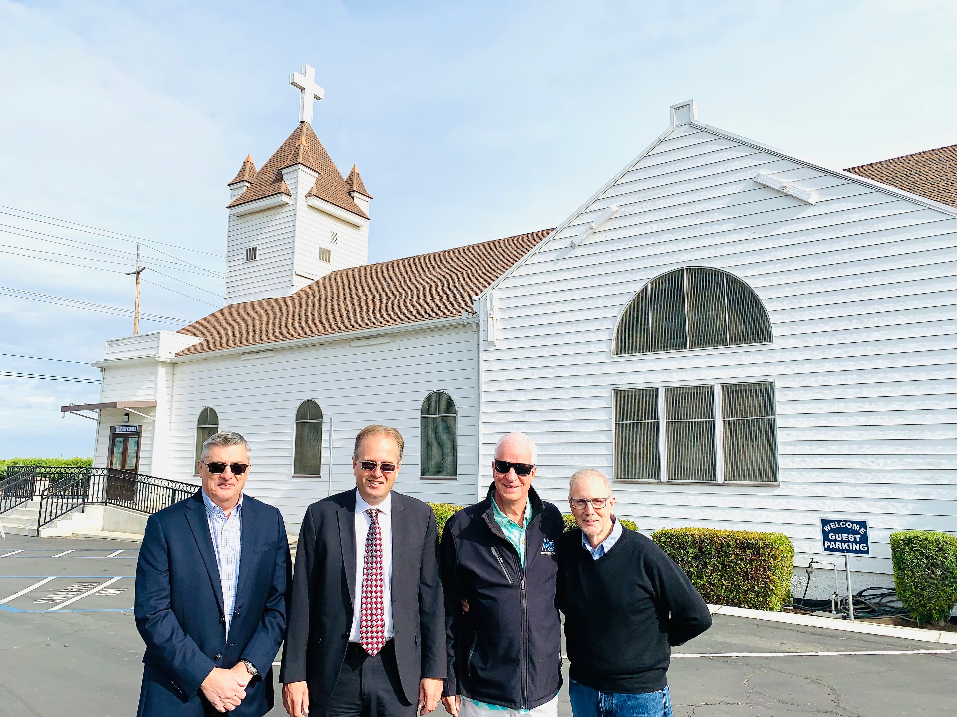 Four men standing in front of a white wooden church with a steeple and cross.