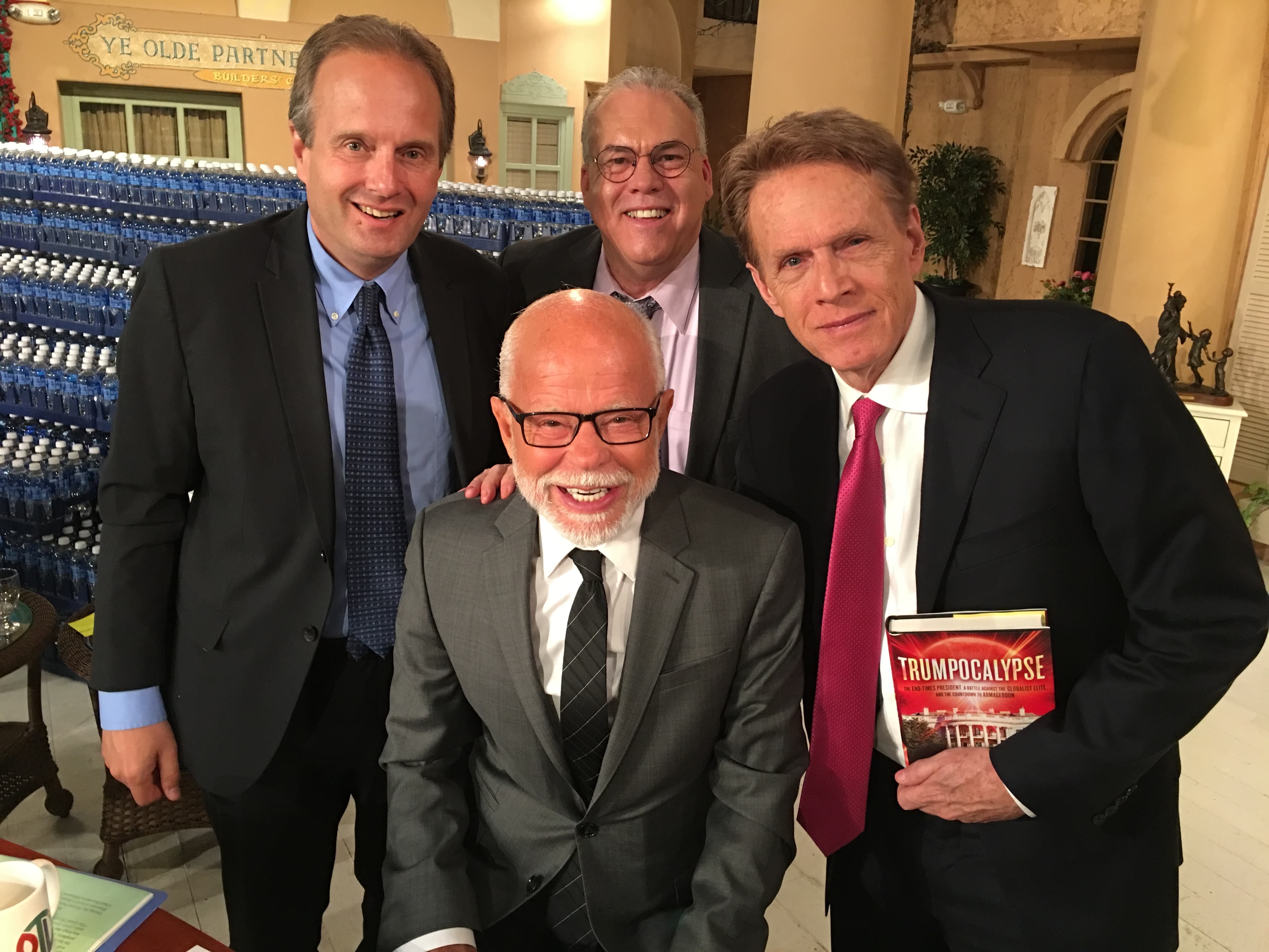 Four men in suits pose together, one holding a book titled "Trumpocalypse" on set.