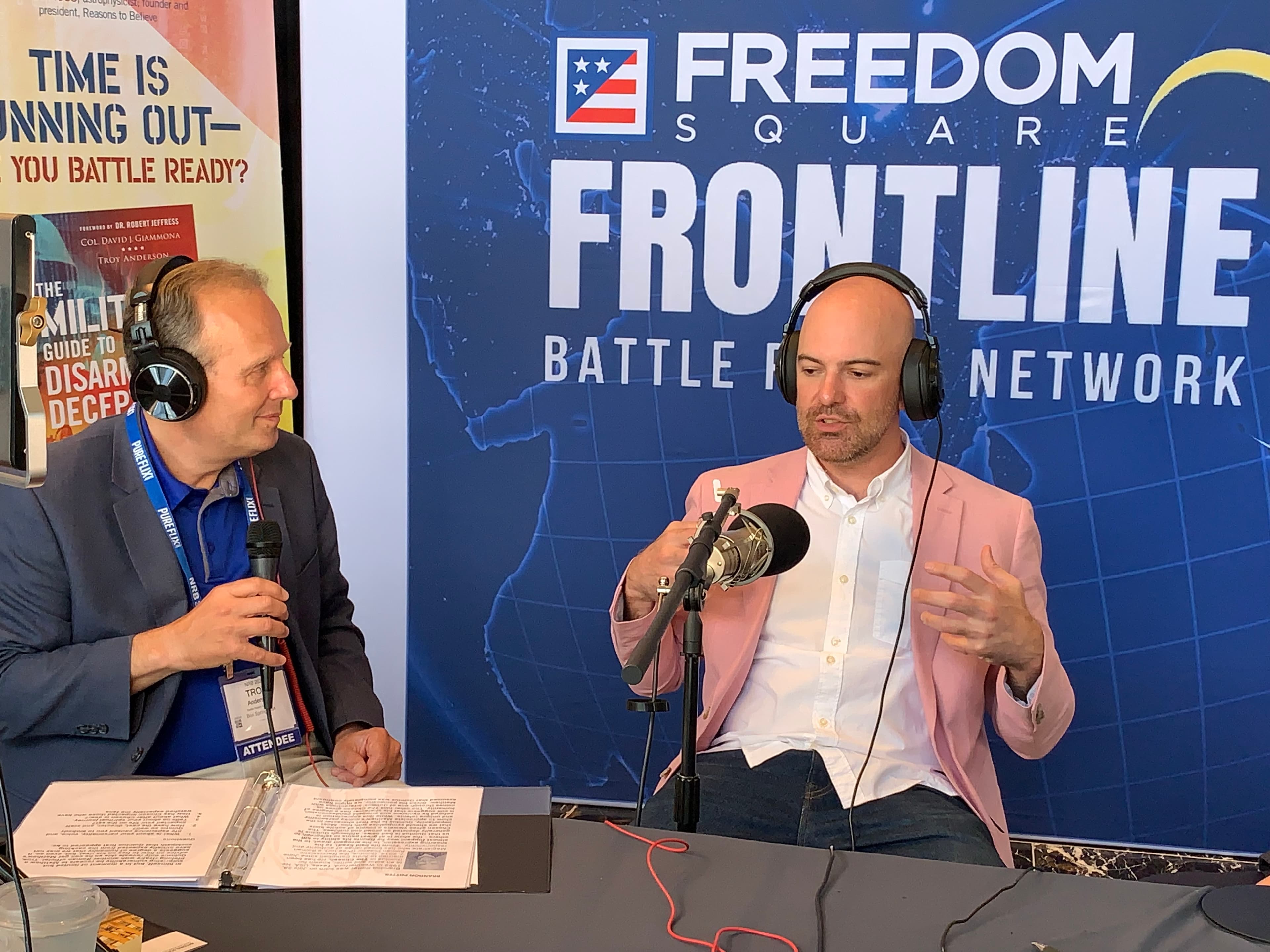 Two men in blazers record a podcast against a Freedom Square Frontline network backdrop.