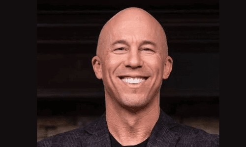 Bald man with a wide smile and white teeth wearing a dark patterned blazer.