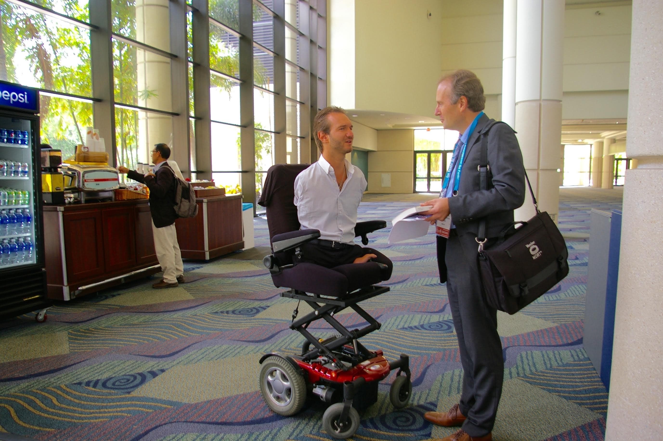 Man without limbs in a motorized wheelchair speaks with a businessman in a lobby.