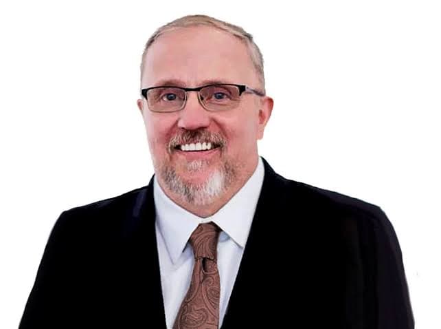 Smiling man with glasses and goatee, wearing a black suit and brown patterned tie.