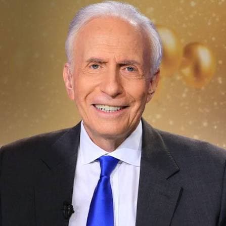 Smiling elderly man with white hair wearing a dark suit and a bright blue tie.