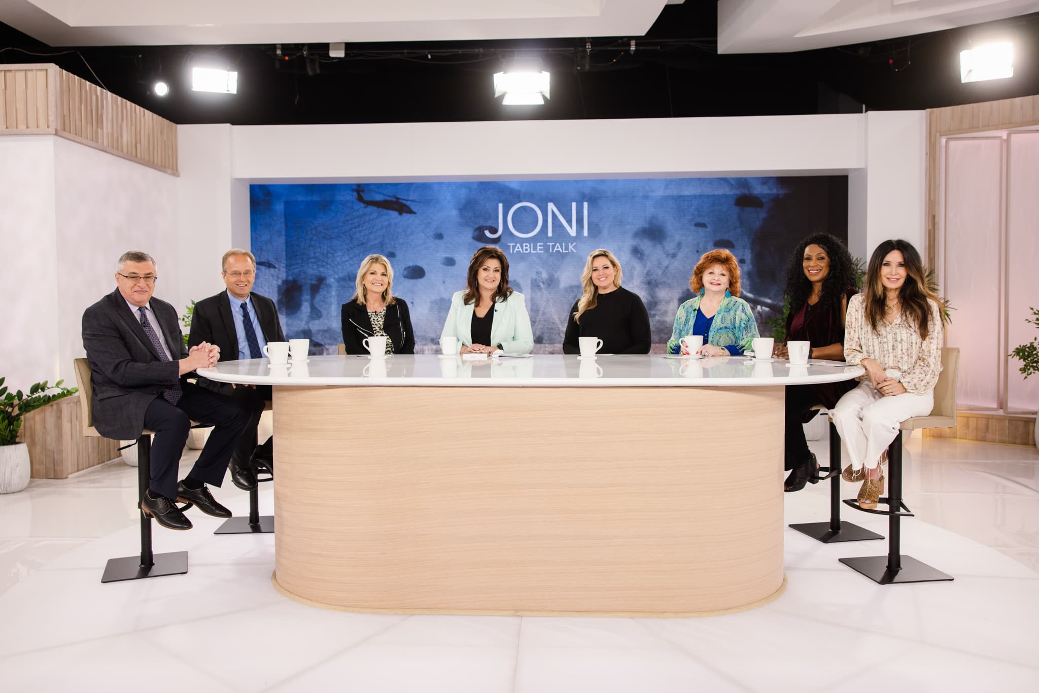 Eight people sit around a large white table on the set of Joni Table Talk.