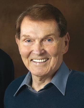 Close-up of a smiling older man with blue eyes, wearing a blue sweater and collared shirt.
