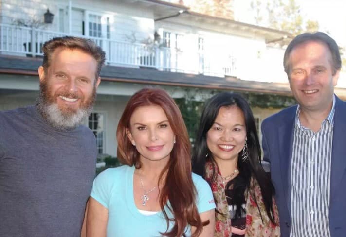 Four smiling people, including a woman with red hair, pose before a large white house.