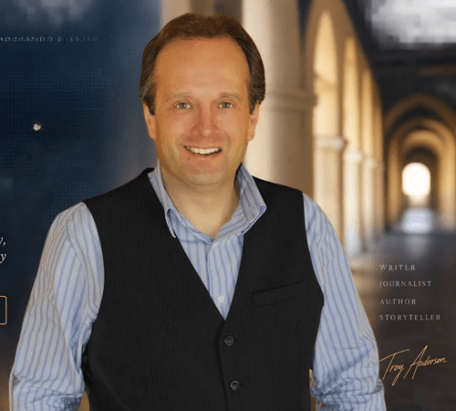 Smiling man in a blue striped shirt and black vest against blurred stone arches.