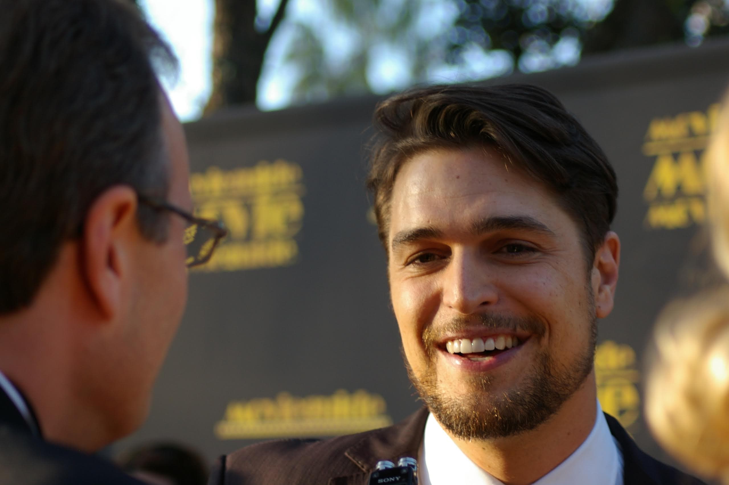 Diogo Morgado smiling during a red carpet interview, wearing a dark suit and white shirt.