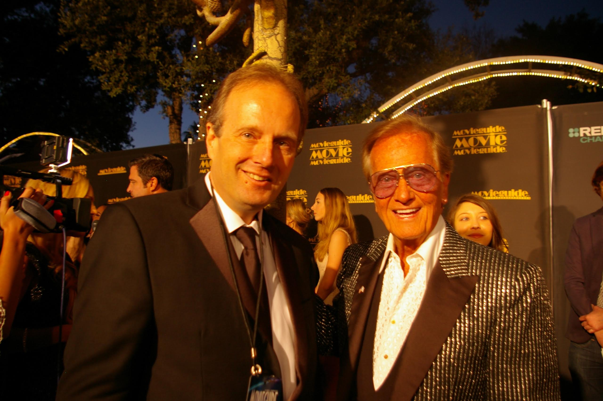 Two men in formal suits smile on a red carpet in front of event signage.
