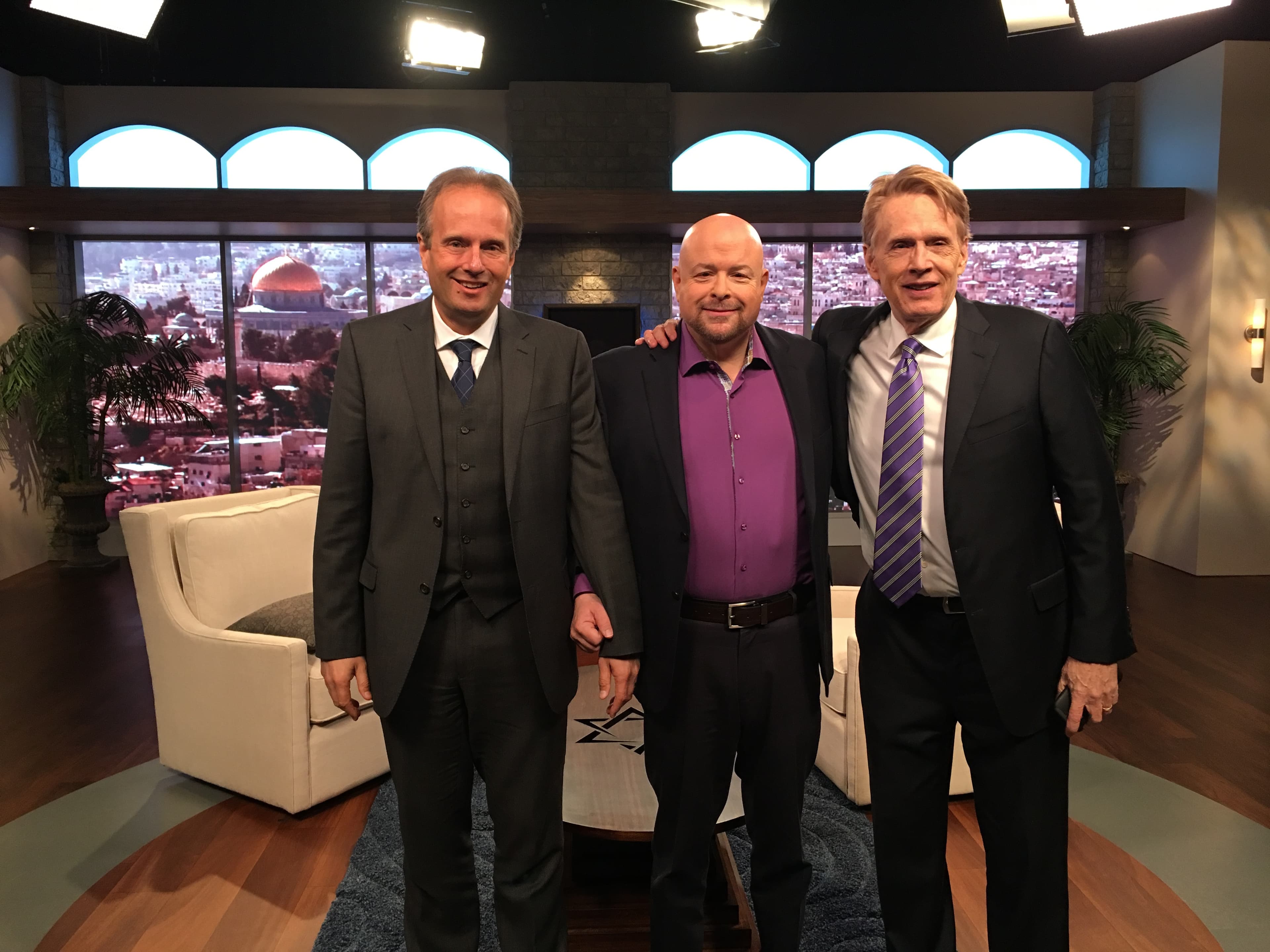Three men in suits pose in a television studio with a Jerusalem cityscape backdrop.
