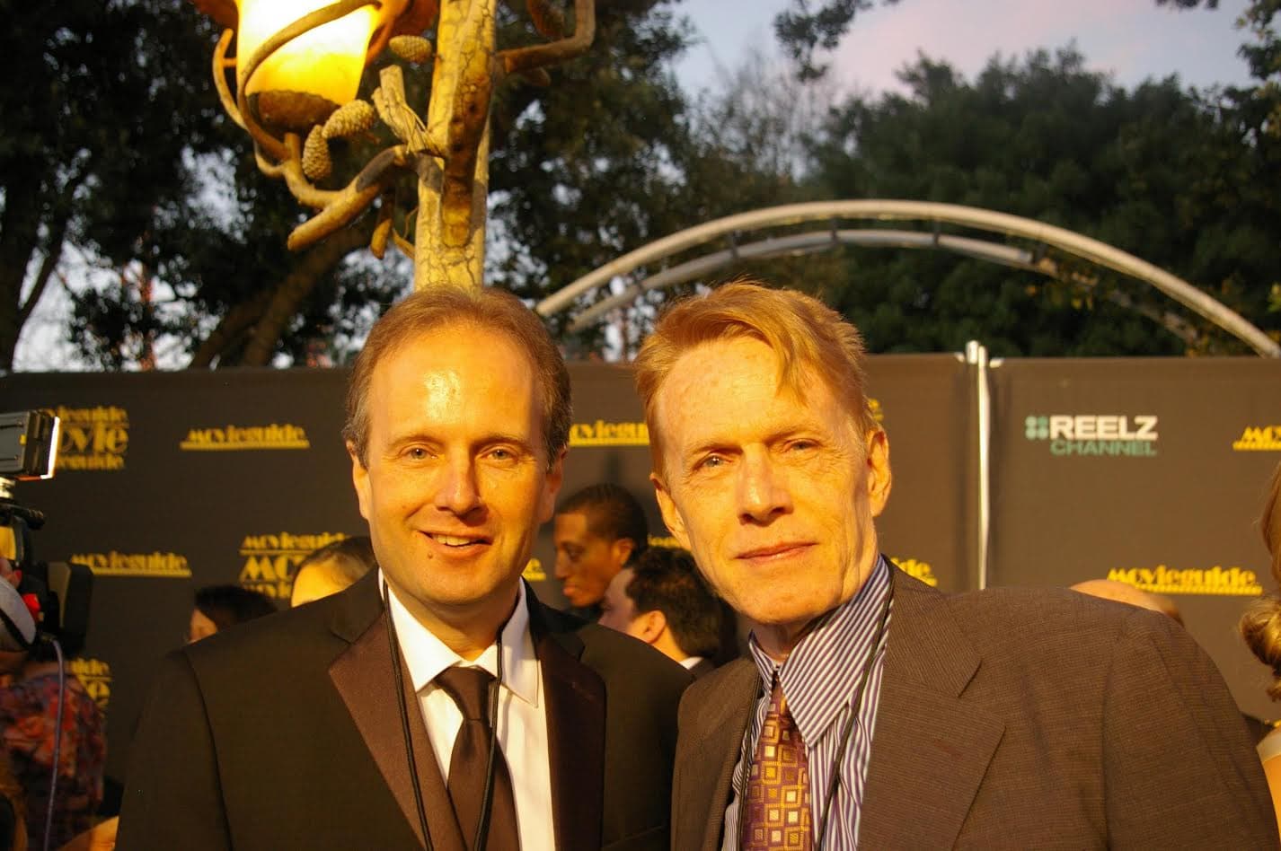 Two men in suits smile at a red carpet event with Movieguide and Reelz logos.